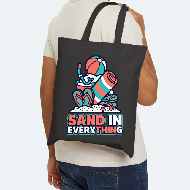 A pile of beach things over SAND IN EVERYTHING, on a black canvas tote