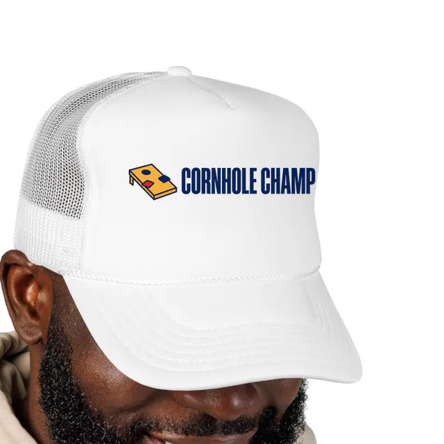 A cornhole board beside CORNHOLE CHAMP, on a white foam trucker hat
