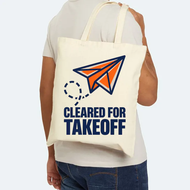A paper airplane climbing on a looping trail, CLEARED FOR TAKEOFF, on a natural canvas tote