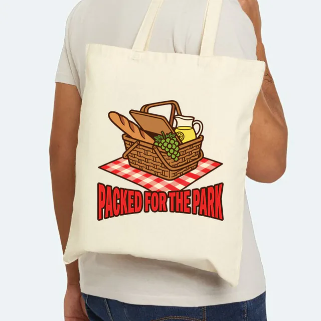 A picnic basket on a gingham blanket, PACKED FOR THE PARK, on a natural canvas tote