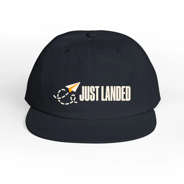 A paper airplane beside JUST LANDED, on a navy surf cap