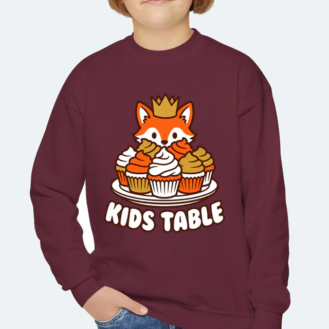 A fox in a paper crown behind the cupcakes, KIDS TABLE, on a maroon youth crewneck