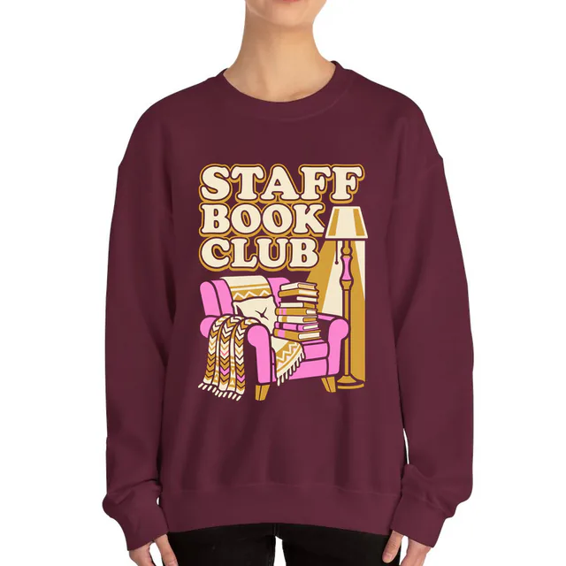 A cozy armchair piled with books for the STAFF BOOK CLUB, on a maroon adult crewneck